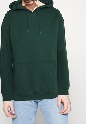 Person trägt einen dunkelgrünen Kapuzenpullover mit Fronttasche, kombiniert mit hellblauen Jeans, vor einem einfarbigen weißen Hintergrund.