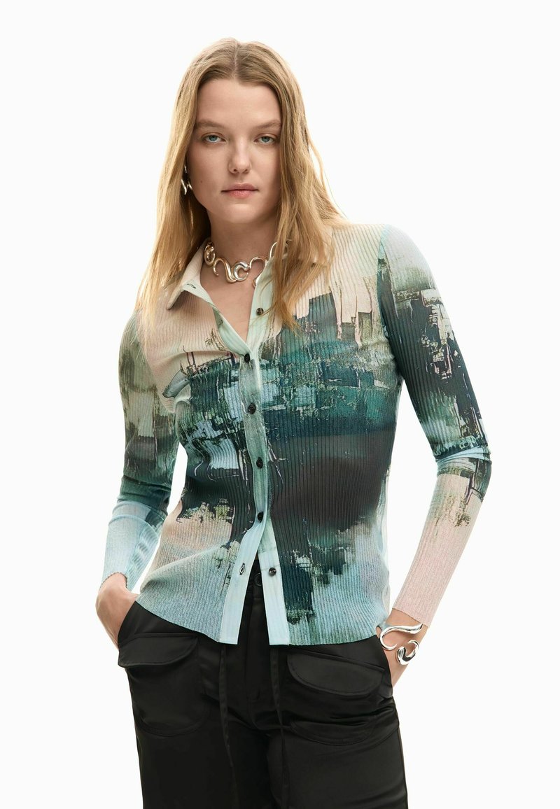 Jeune femme aux longs cheveux blonds portant une chemise côtelée à boutons à motifs abstraits verts et beiges, et un pantalon noir, les mains dans les poches.