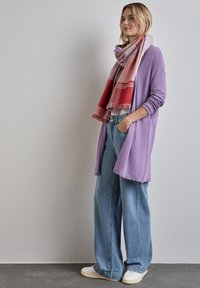 Cardigan lavande clair, coupe ample, porté sur une chemise blanche ; associé à un jean bleu ample et une écharpe à carreaux rouge et rose. Baskets blanches.
