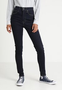 Jeans escuros de cintura alta com um corte Slim e costura em contraste, combinados com sapatilhas baixas azul-marinho e uma camisa de botão clara com riscas.
