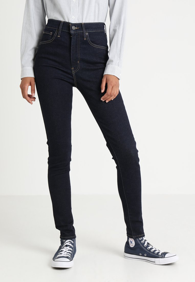 Jeans escuros de cintura alta com um corte Slim e costura em contraste, combinados com sapatilhas baixas azul-marinho e uma camisa de botão clara com riscas.