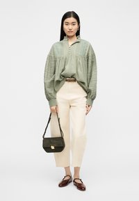 Γυναίκα που φοράει ανοιχτοπράσινη κεντημένη μπλούζα με φουσκωτά μανίκια, κρεμ παντελόνι, σκούρα καφέ παπούτσια και κρατάει μια μικρή σκουροπράσινη τσάντα.