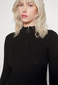 Pull en maille noir côtelée avec un col montant et un détail de fermeture éclair argentée. Présente des manches longues et une coupe ajustée. Texture en maille légère.