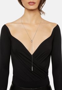 Long silver necklace featuring a circular pendant and a straight bar, worn over a black deep-V neck top with gathered fabric details.