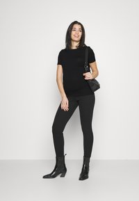 Tricou negru, ajustat, asortat cu colanți negri. Botine negre cu toc pătrat. Geantă mică neagră tip crossbody. Fundal simplu.