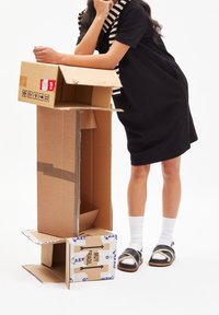 Tekturowe pudełka w różnych rozmiarach, brązowe z etykietami wysyłkowymi. Osoba nosi czarną sukienkę i wciągnięty szal w paski, stoi w sandałach i białych skarpetkach.