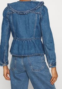 Denim jacket with a peplum waist, ruffled collar, and button accents. Dark blue fabric, with a slightly faded texture and fitted shoulders.