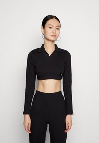 Top preto cropped de mangas longas com decote colarinho, textura canelada e pequeno detalhe da marca perto da bainha. Combinado com calças pretas de cintura alta.