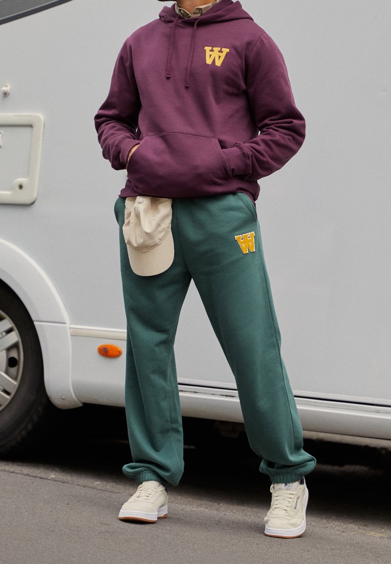 Vínová mikina s žlutým logem, zelené tepláky a krémové tenisky. Součástí je lehká čepice na kalhotách, které mají volný střih a měkkou texturu.