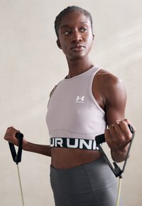 Fitt nő sporttopban és leggingsben ellenállási szalaggal edz, oldalra koncentrálva egy sima háttér előtt.