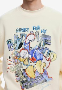 Donald Duck en tenue de marin bleue avec le poing levé, avec le texte « Désolé pour ma mauvaise humeur » et un fond de style bande dessinée sur un sweat-shirt crème.