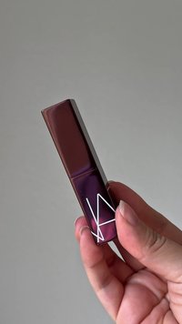 LippenGloss in einer rechteckigen Tube mit glänzender dunkelburgunderfarbener Oberfläche und einem weißen geometrischen Logo, das auf der Vorderseite gedruckt ist.