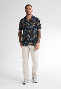 Camicetta blu navy a maniche corte con stampa di palme multicolore, abbinata a pantaloni beige chiaro e scarpe da ginnastica bianche.