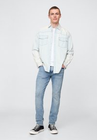 Világoskék farmering két mellzsebbel, világoskék farmernadrággal és fekete magas szárú tornacipővel párosítva. Semleges háttér.