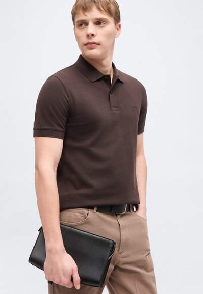 Jeune homme portant un polo marron foncé et un pantalon beige, tenant une pochette en cuir noir sur un fond clair uni.