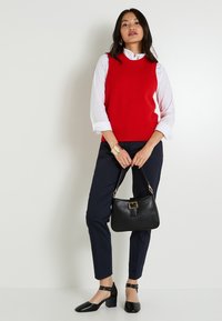Mujer que lleva un suéter sin mangas rojo sobre una camisa blanca, pantalones marinos, tacones negros, sosteniendo un bolso negro frente a un fondo blanco.