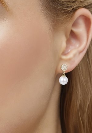 Close-up van een oor met een gouden oorbel met kleine diamanten en een hangende witte parel op een lichte huid en blond haar.