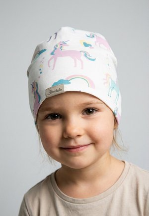 Young child wearing a white hat with pastel unicorns and rainbows, smiling gently against a plain light background.