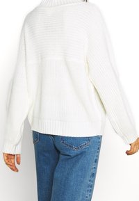 Vit stickad tröja med hög krage, ribbade muddar och nederkant. Har en lös passform och texturerat mönster. Matchad med blå jeans.