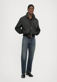 Black bomber jacket with a high collar, front zip, and two large pockets, paired with blue denim jeans and black leather shoes.