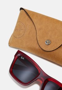 Ray-Ban solbriller med røde transparente rammer og grå linser, ledsaget af et brunt læderetui med logo og tryklås.