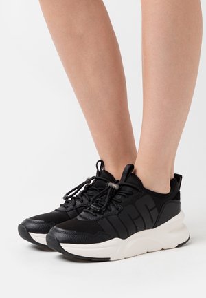 Chaussures de sport noires avec un dessus texturé, un logo audacieux, un design léger et une semelle de couleur crème contrastante.
