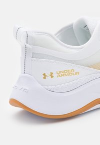 Vit träningsskor med mesh och syntetiska material. Har en texturerad sula, guldlogotyp och kontrasterande orange accent på yttersulan.