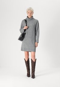 Robe turtleneck en tricot gris avec un motif texturé, portée avec des bottes de cowboy en cuir marron ornées de coutures décoratives. Sac à main noir porté.