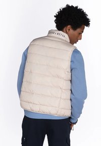 Chaleco acolchado beige con cuello alto, con costuras horizontales y un logo en el cuello. Llevado sobre una camisa de manga larga azul claro.