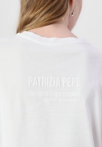 Bela bombažna majica s krojačkim ovratnikom. Ima reliefno bele napise na hrbtni strani, vključno z imenom blagovne znamke "Patrizia Pepe" in dodatnimi frazami.