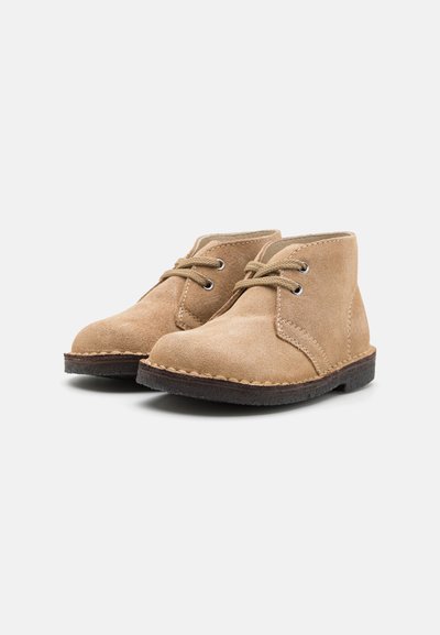 Botine bej din suede cu un design cu vârf rotund, închidere cu șireturi și o talpă din cauciuc maro. Prezintă accente de cusătură de-a lungul marginii.