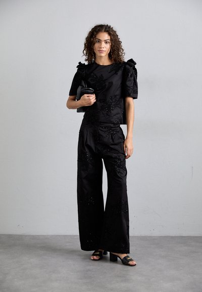 Juodas dekoruotas dviejų dalių kostiumas, kuris apima trumpas rankoves turintį viršų su pūstomis petnešomis ir plačiąsias kelnes su gėlių motyvais.