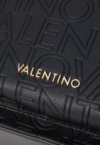 Crna kožna torba s reljefnim uzorkom logotipa i zlatnom natpisom "VALENTINO", glatke teksture i strukturiranog oblika.