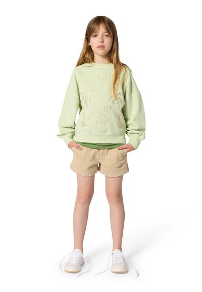 Jeune fille debout, les mains dans les poches, portant un sweat-shirt vert clair avec des palmiers, un short beige et des baskets blanches défaites.