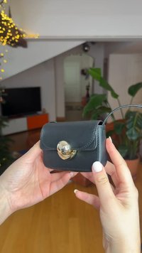 Sac à main en cuir noir avec un fermoir rond en or, design compact, texture lisse et poignée supérieure pour le porter.