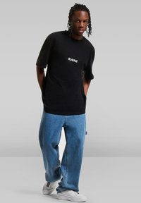 Black short-sleeve T-shirt with "KANI" logo, paired with loose blue denim jeans and white sneakers. Smooth fabric and relaxed fit.