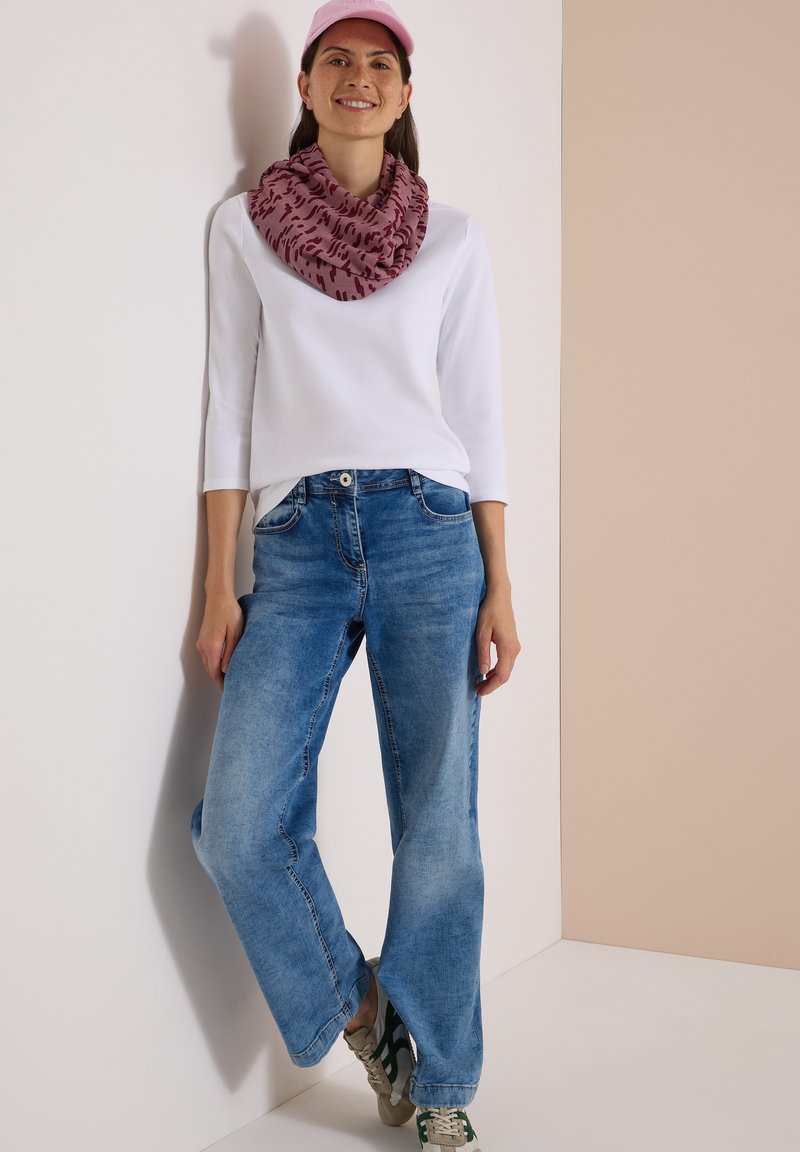Femme portant une casquette rose, une écharpe violette à motifs, un haut blanc à manches longues, un jean bleu ample et des baskets, debout contre un mur bicolore.