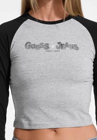 Gray cropped shirt with black raglan sleeves. Features "Guess Jeans" logo in textured print, and the phrase "since 1981." Soft cotton material.