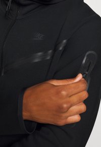 Svart Nike-hoodie med dragkedja, med en blank rand över bröstet och en texturerad ficka på armen. Mjukt, stretchigt tyg.