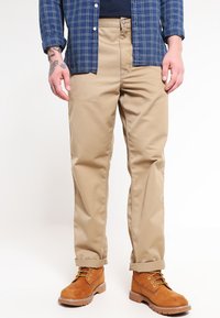 Beige rechte-stijl broeken van stevige stof, met een eenvoudig ontwerp en voorzakken, gecombineerd met bruin veterboots en een blauw geruit shirt.