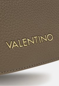 Hnědá texturovaná kožená taška má zlaté logo "Valentino". Švy jsou viditelné podél okraje, což zdůrazňuje její designové detaily.