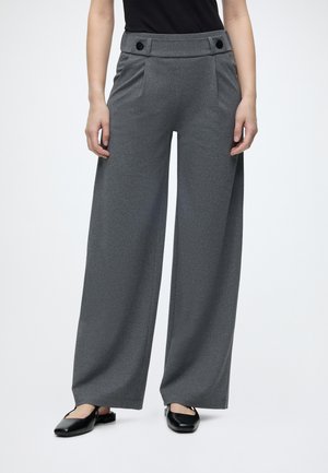 Personne portant un pantalon gris taille haute à jambes larges avec des détails de boutons à la taille et des chaussures plates noires à bout carré.