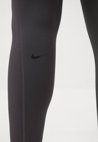 Czarne legginsy sportowe mają gładką teksturę, dopasowany krój, widoczne szwy oraz małe logo Nike w kolorze czarnym na udzie.
