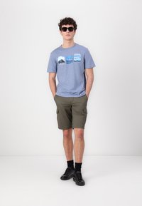 Blå bomull T-skjorte med grafiske trykk; olivenfargede cargo shorts; svarte sneakers; solbriller. Modellen står med hendene i lommene, nøytral bakgrunn.