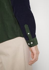 Långärmad skjorta med en grön kordflortskropp och marinblå ärmar. Inkluderar en grön kordflortsmudd med vita knappar och en spetsig design.