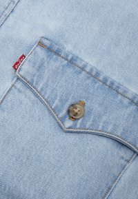 Light blue denim fabric featuring a pocket with a plastic button and contrasting orange stitching. Label visible in the top left corner.