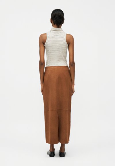 Γυναίκα με σκούρα μαλλιά σε κότσο, που φοράει αμάνικη λευκή και γκρι ριγέ μπλούζα, καφετιά midi φούστα ψηλόμεση και μαύρα παπούτσια slip-on, με την πλάτη στραμμένη.