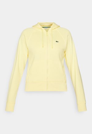 Sudadera amarilla con cremallera que cuenta con una capucha, mangas largas tipo raglán, un bolsillo frontal y un pequeño logo verde en el área del pecho izquierdo. Material suave.