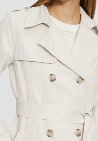 Beżowy trencz z podwójnym rzędem guzików, dużymi guzikami, paskiem w talii i klasycznym kołnierzem. Wykonany z gładkiej tkaniny.