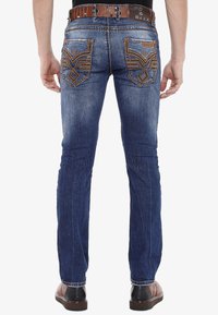 Jean en denim bleu foncé, coupe slim avec broderies contrastantes sur les coutures des poches, cinq poches et ceinture en cuir marron.